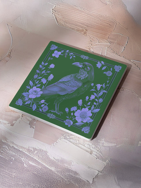 Square ceramic coaster featuring intricate lavender illustration of a raven surrounded by botanical elements on forest green background, with ornate border design and cork backing.